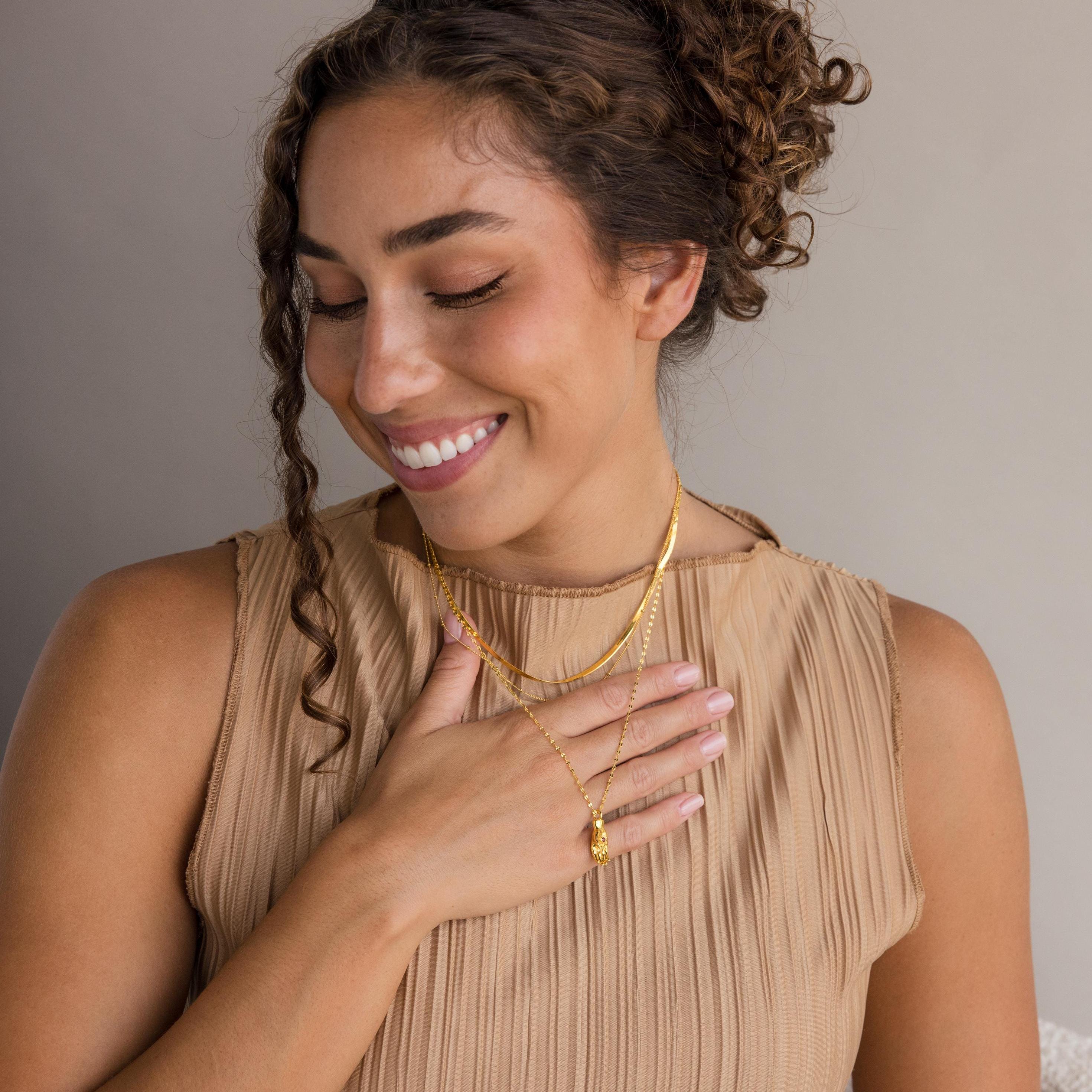 A smiling woman with curly hair wears gold necklaces, including the Evil Eye Birthstone Hand Necklace, paired with a beige pleated top and her hand resting on her chest.