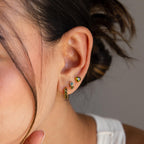 A woman's ear adorned with three gold and gemstone earrings, including the Toi et Moi Birthstone Studs. With her hair tied back and a white top, these studs are shown as an ideal personalized earring gift.