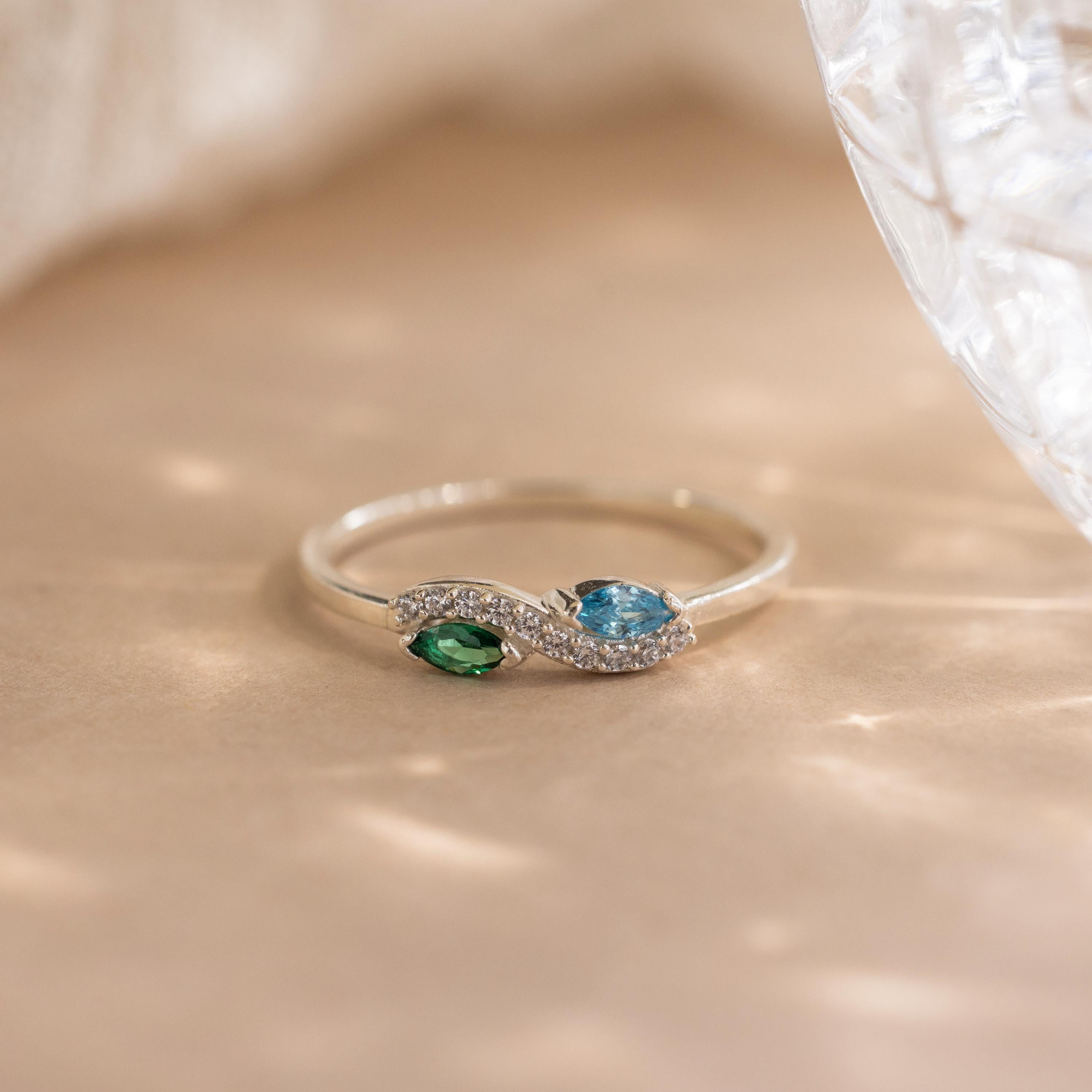 The Infinity Marquise Birthstone Ring in silver features green and blue gemstones with clear accents, elegantly displayed on a soft beige surface. This customizable ring showcases a timeless infinity design.