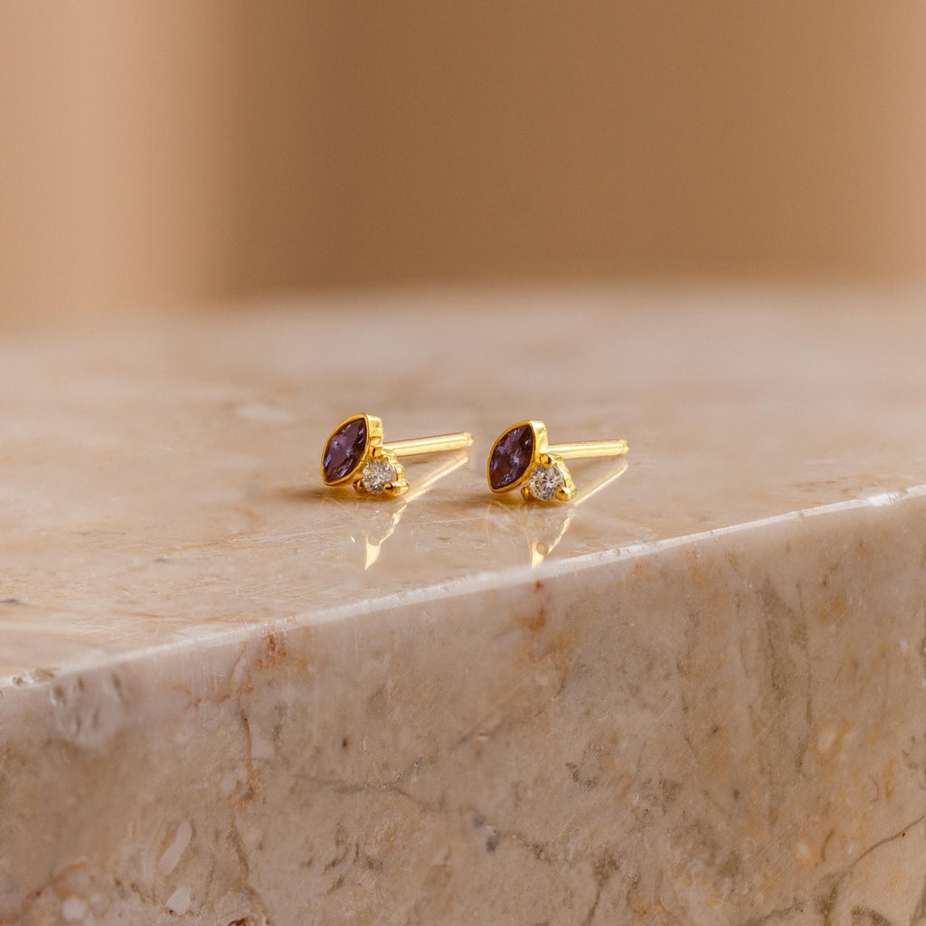 Diamond Marquise Birthstone Studs featuring purple and clear gemstones are displayed on polished marble. These marquise-cut earrings bring an elegant touch to any look.