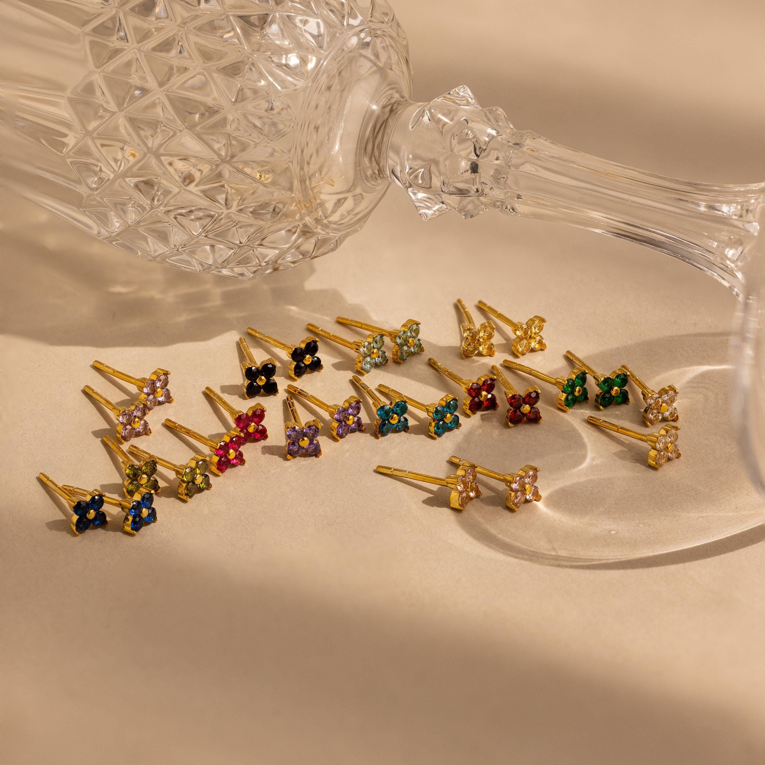 An Ivy Flower Birthstone Stud lies beside a crystal glass on its side, with gold and sterling silver stud earrings elegantly arranged on a beige surface.