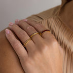 A hand with neatly manicured nails wears the Engraved Birthstone Eternity Ring and two gold bands, resting gracefully on beige fabric.