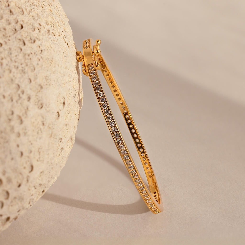Diamond Tennis Bangle with a sleek, curved design and embedded sparkling stones, displayed leaning against a textured beige rock on a neutral background.