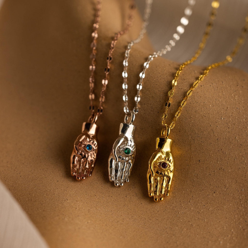 Three metallic Evil Eye Birthstone Hand Necklaces—rose gold, silver, and gold—each with a gem, rest on a textured background. This unique collection blends elegance with spiritual significance.