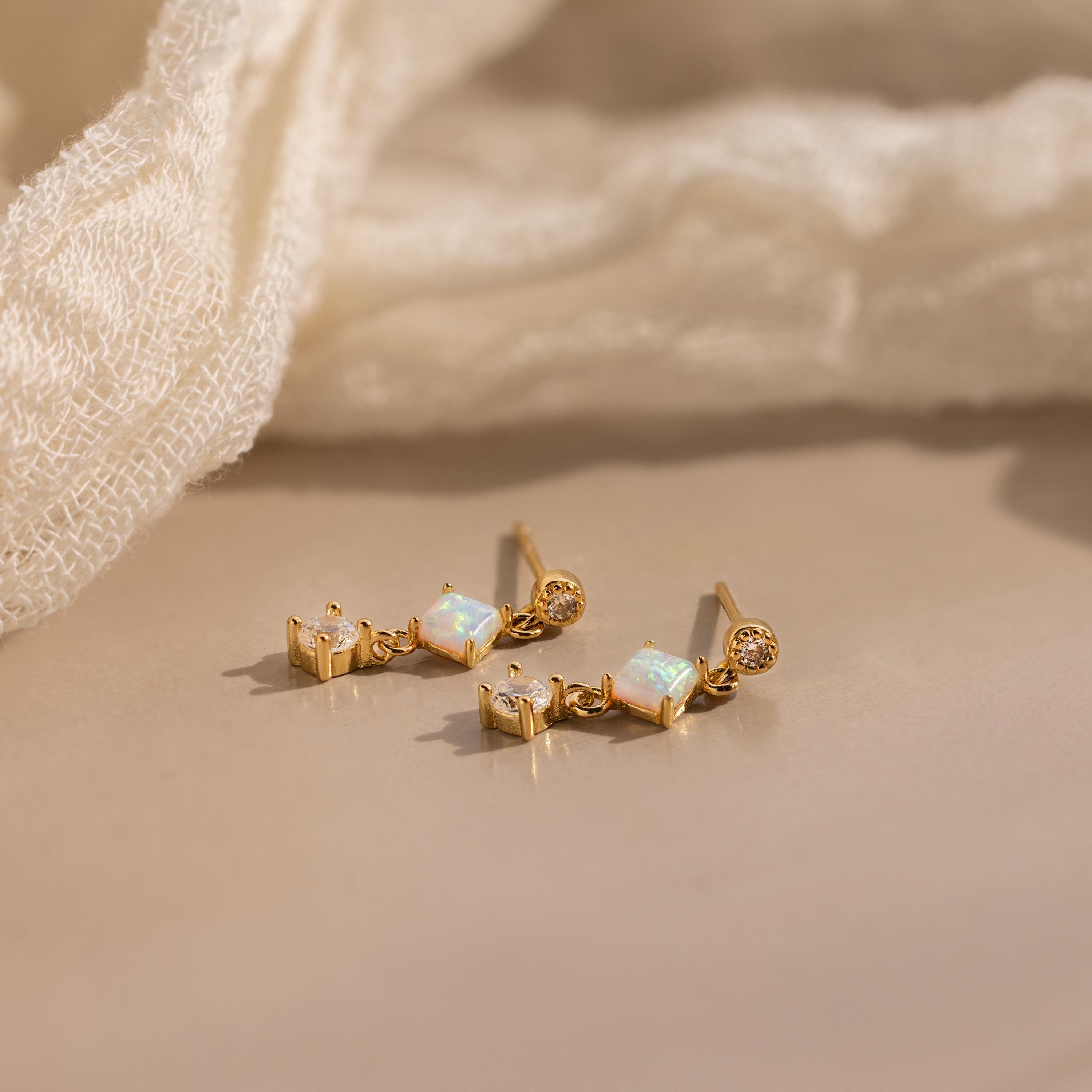 The Opal & Diamond Drop Studs, featuring opal and sparkling diamonds, rest on a beige surface beside a soft fabric—perfect dainty earrings for any occasion.