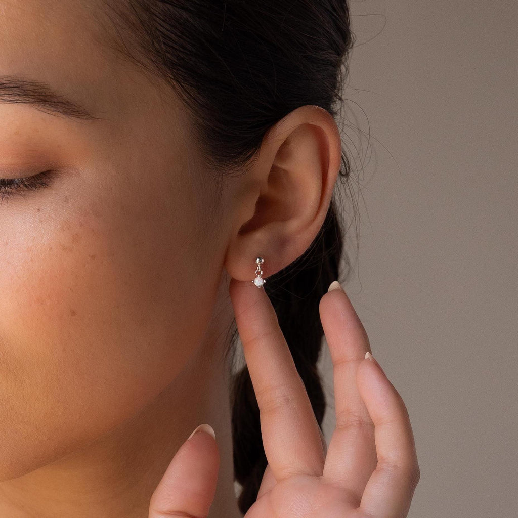 A person with braided hair gently touches a Dainty Opal Drop Stud, admiring the earring's delicate silver shine.