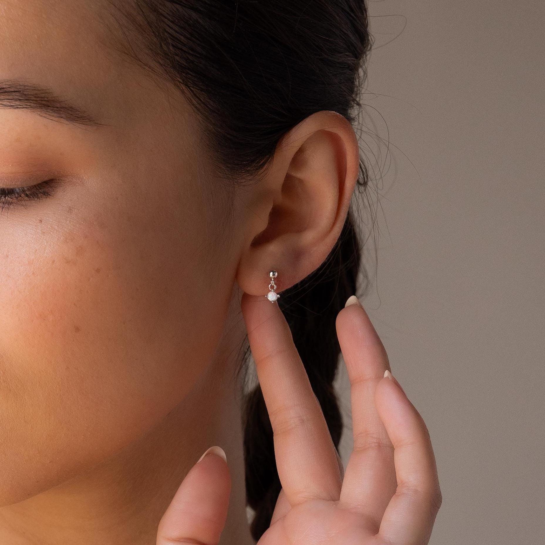 A person with braided hair gently touches a Dainty Opal Drop Stud, admiring the earring's delicate silver shine.
