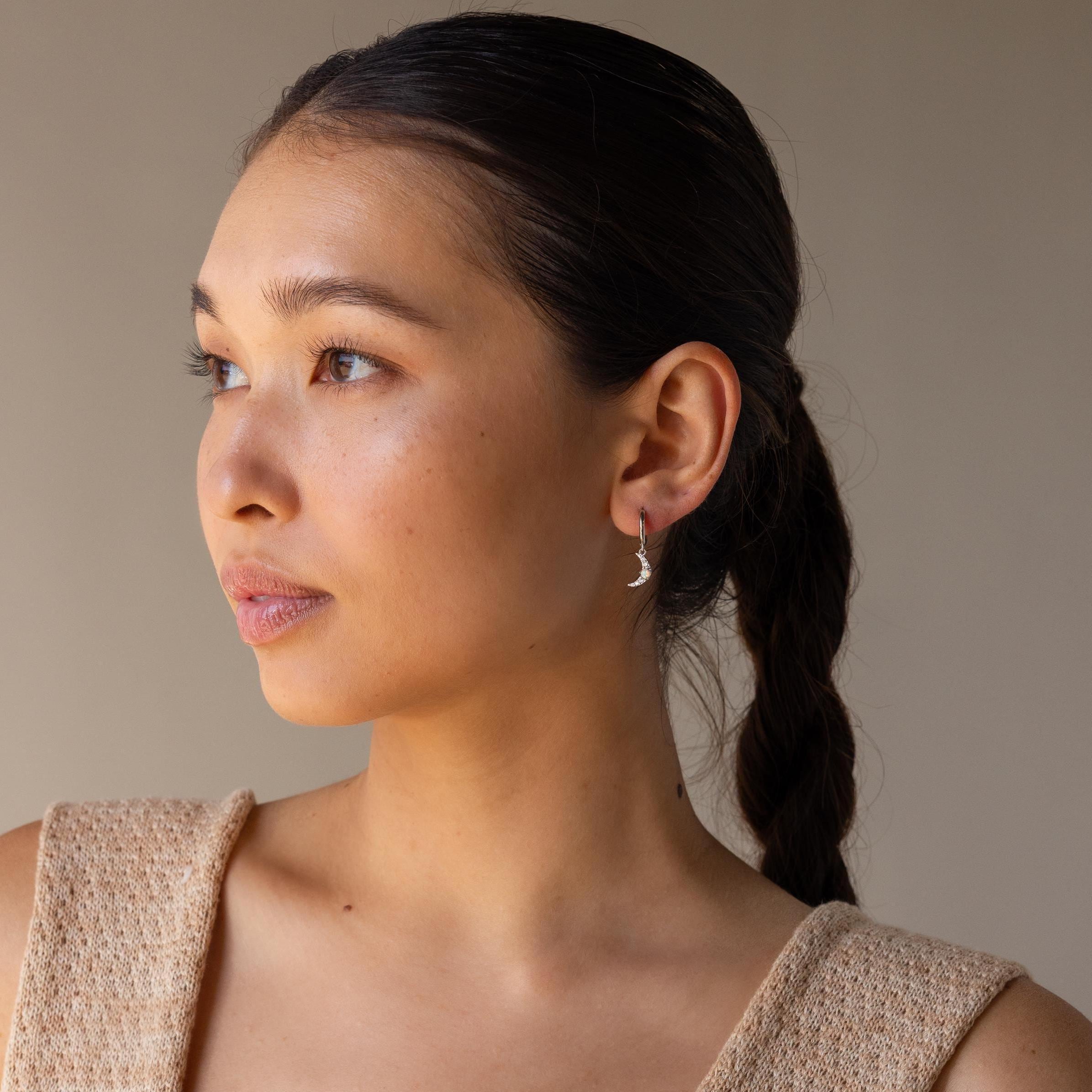 A woman with braided hair wears a beige sleeveless top and Opal Sun & Moon Huggies, featuring delicate crescent moon details, as she looks to the side.