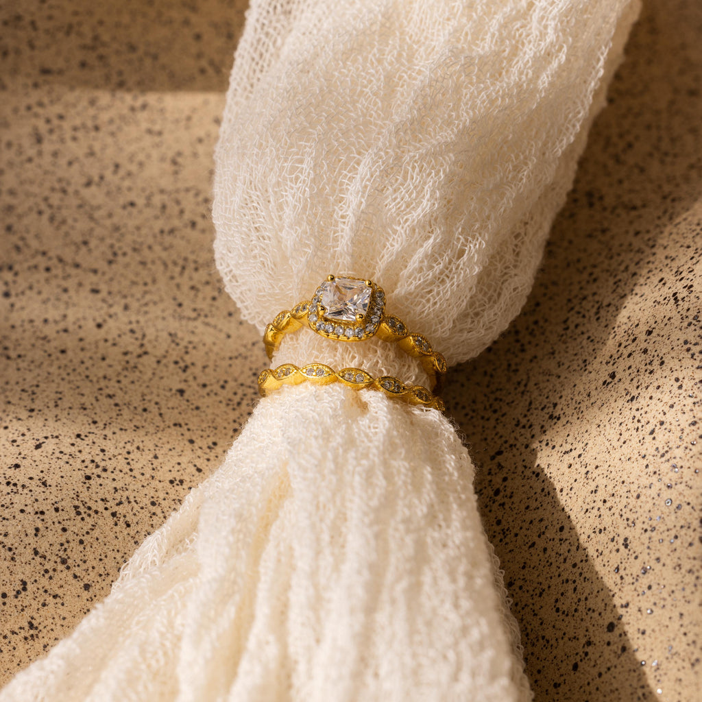 The Vintage Diamond Rings Set, featuring a cushion-cut halo ring and a marquise-detailed band, rests on textured cream fabric in the sunlight.