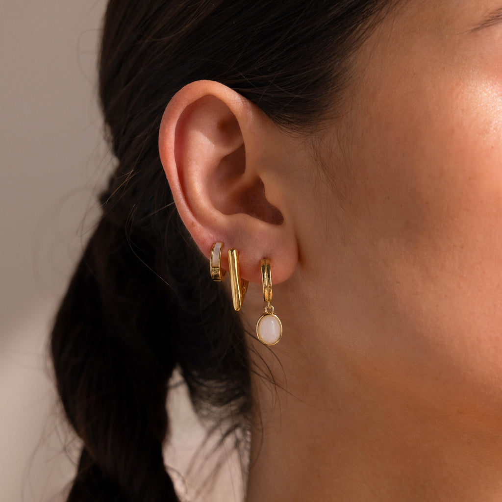 Close-up of a woman's ear adorned with three gold earrings, including Moonstone Drop Hoops featuring a white circular stone.