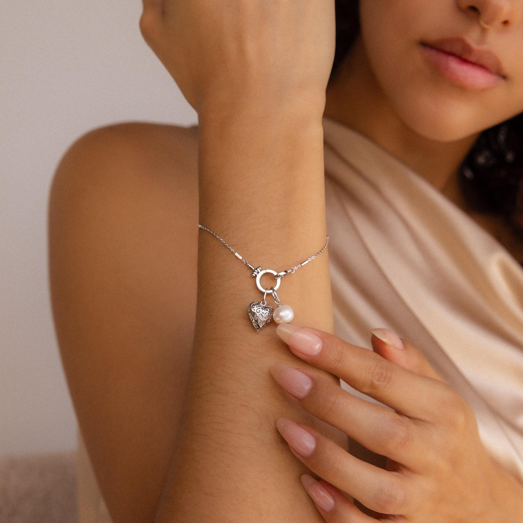 A woman wears the Hinged Circle Charm Bracelet in Sterling Silver, delicately touching the minimal heart and pearl charms featured on this elegant jewelry piece.