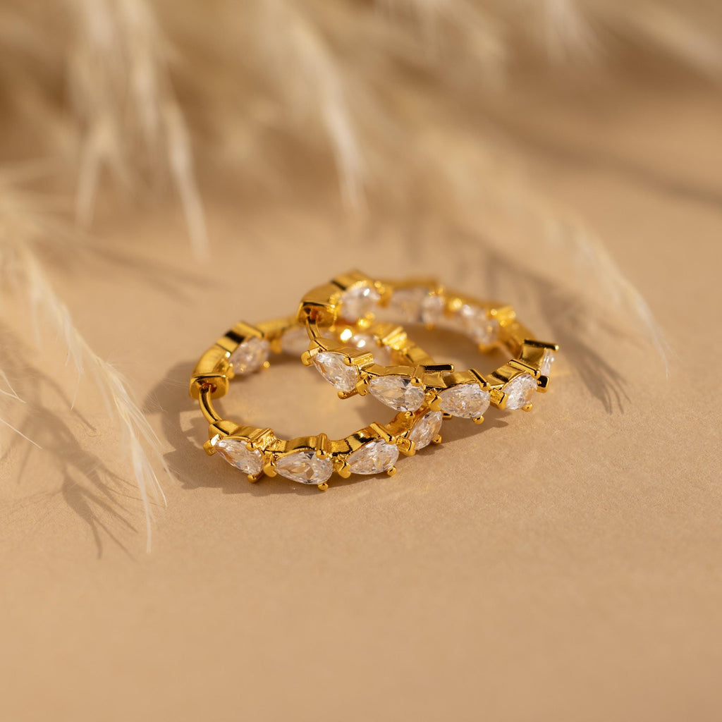 The Inside Out Pear Diamond Hoops, gold statement earrings featuring marquise-cut clear stones, rest elegantly on a beige surface with a blurred plant behind—an ideal bridesmaid gift.