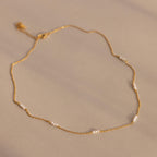 The Hali Pearl Station Necklace features delicate gold links with small white pearls, shown on a neutral beige background—ideal for those who love minimalist jewelry.