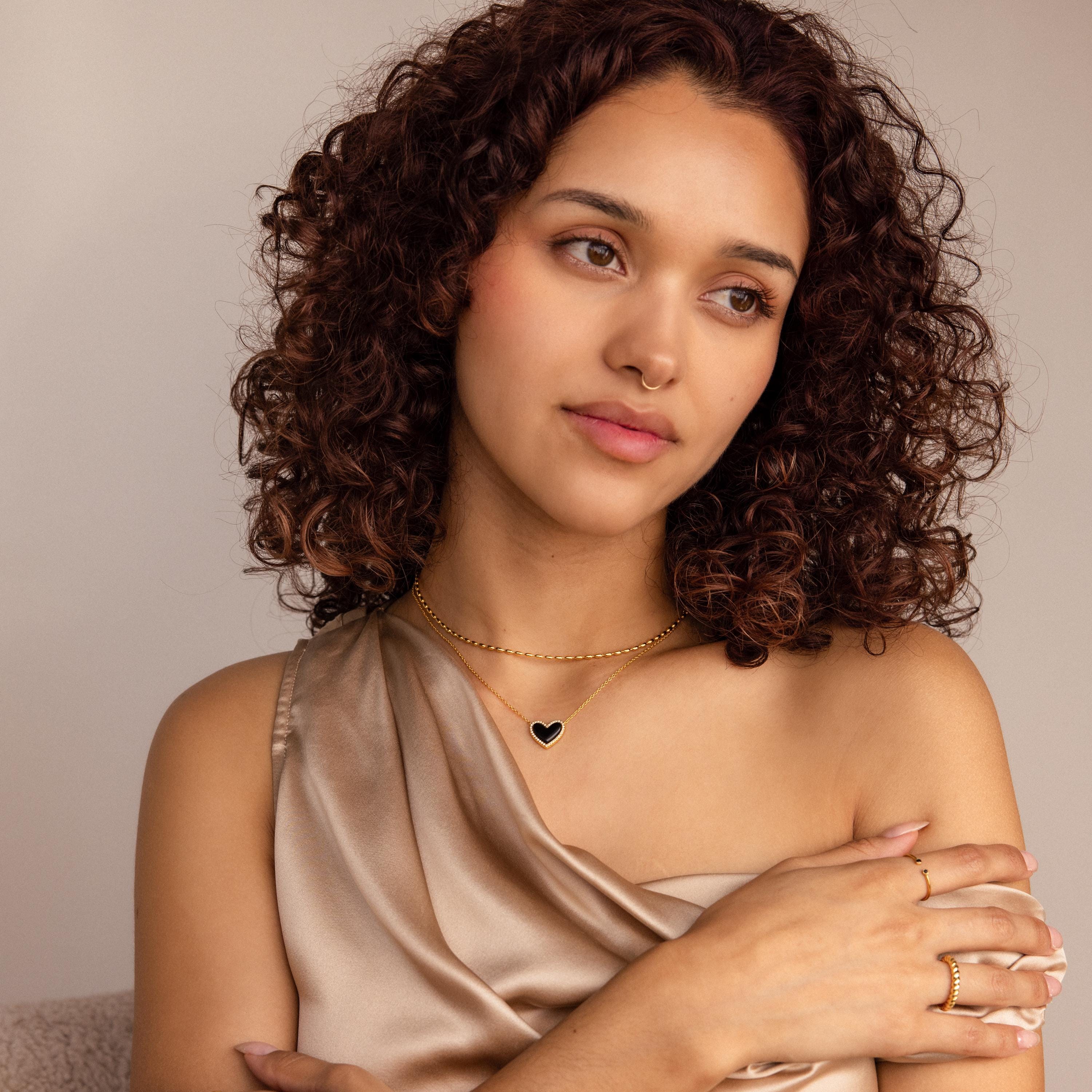 A woman with curly hair wearing a satin top and the Oval Beaded Necklace, layered with a gold heart pendant and rings, gazes thoughtfully to the side.