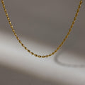 The Oval Beaded Necklace features a simple gold design with a twisted chain, displayed against a gray background and casting a soft shadow.
