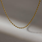 The Oval Beaded Necklace features a simple gold design with a twisted chain, displayed against a gray background and casting a soft shadow.