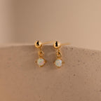 Dainty Opal Drop Studs showcase small round opal gemstone dangles on gold, displayed on a textured beige surface. Lightweight and elegant, these earrings are perfect for effortless style.