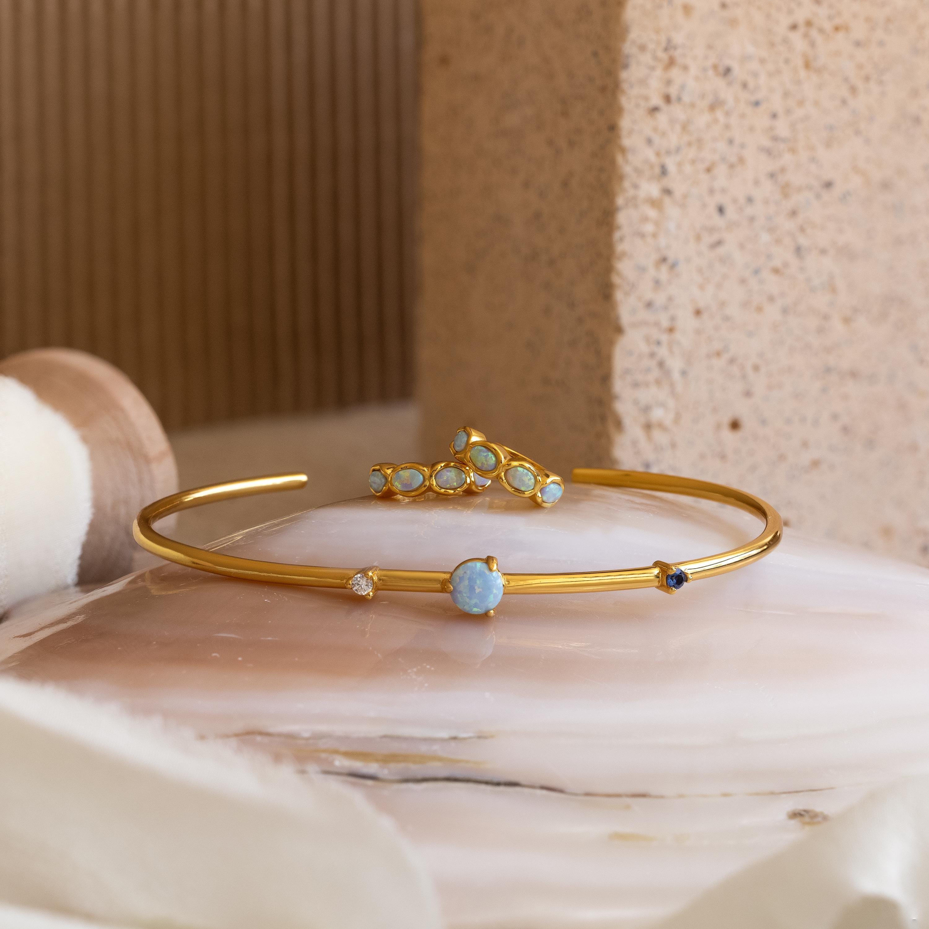 Blue Opal Beaded Huggies paired with gold bangles adorned with opal and blue gemstones, displayed on a polished stone surface against a neutral background.