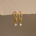 The Dainty Opal Drop Huggies feature gold hoops with small round opal charms and rest on a textured beige surface, creating delicate earrings that add a touch of elegance to your everyday style.