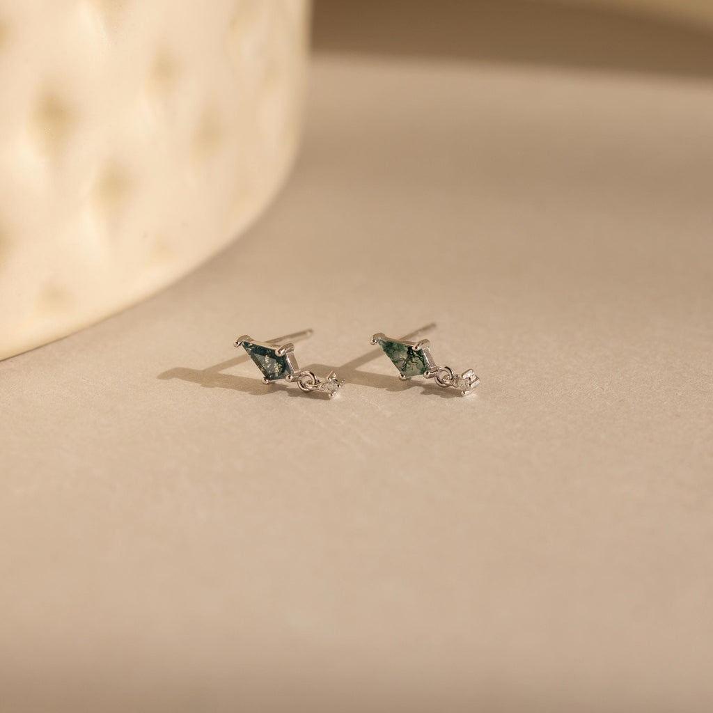 A pair of Agate Kite Drop Earrings with small silver studs and moss agate stones rests on a softly lit beige surface.