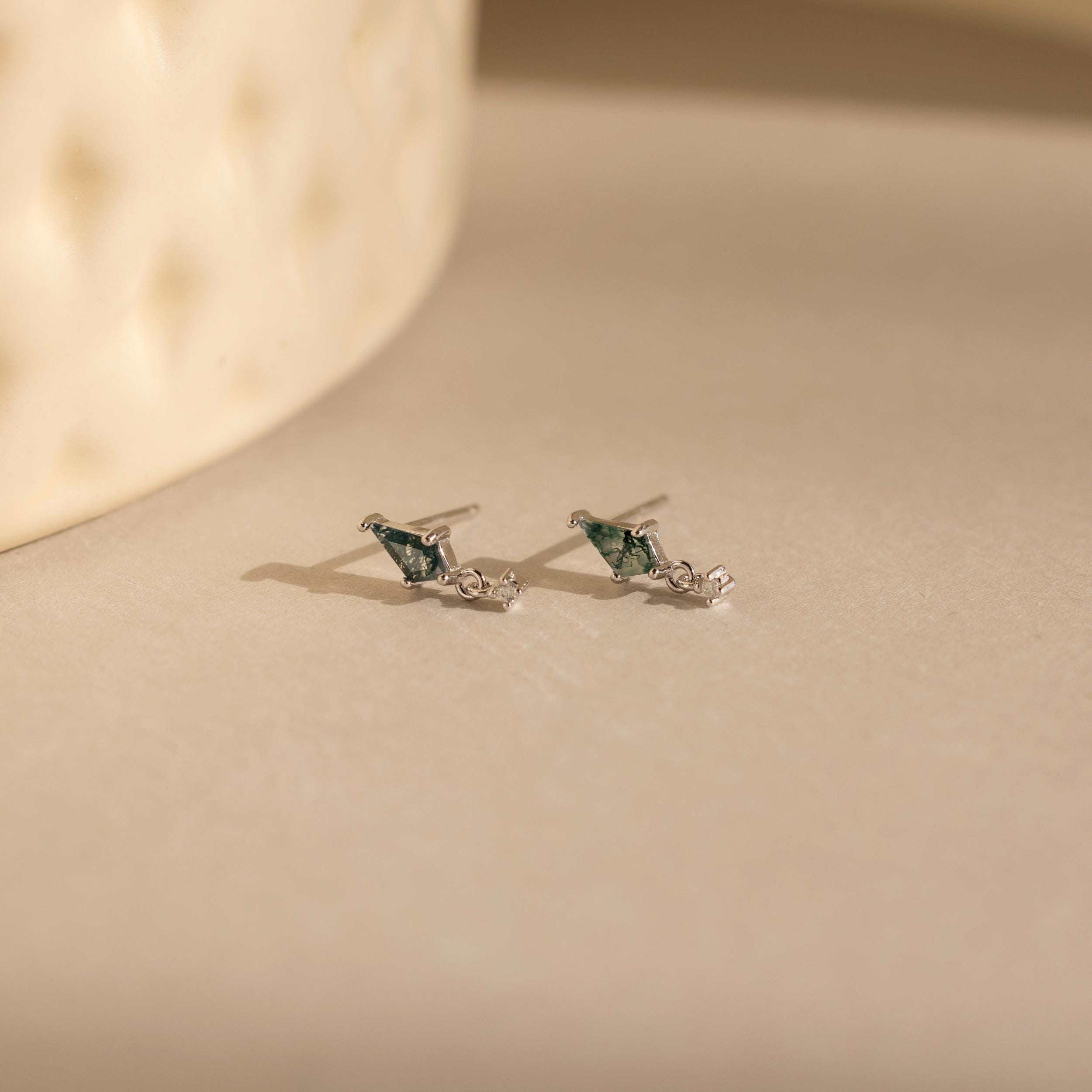 A pair of Agate Kite Drop Earrings with small silver studs and moss agate stones rests on a softly lit beige surface.