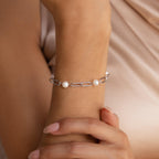 A woman’s wrist showcases the Pearl Station Paperclip Bracelet—silver chain with three pearls—set against a soft beige background.