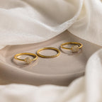 The Classic Pave Ring Set, featuring three gold rings with diamond accents, is elegantly displayed on a beige surface with soft white fabric—an ideal jewelry set for any occasion.
