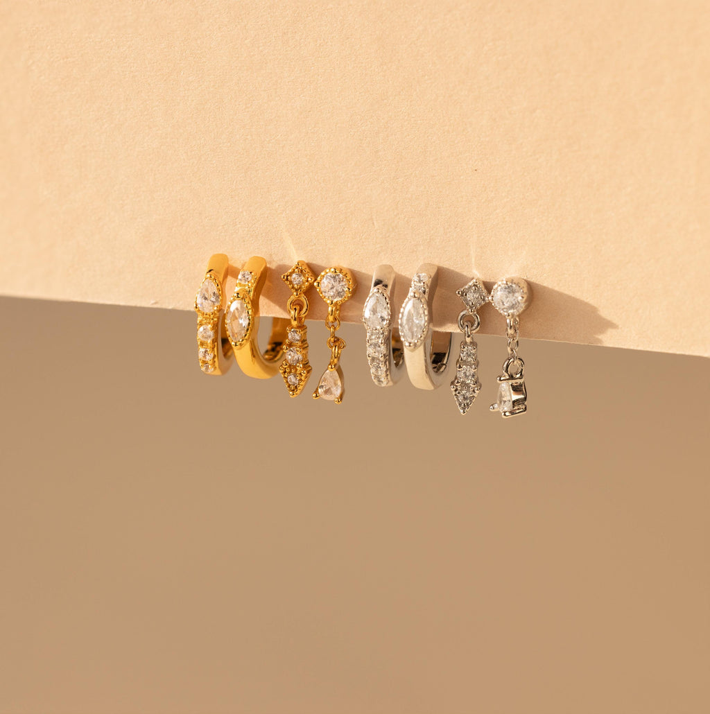 The Dainty Pave Earrings Set, featuring five pairs of gold and silver hoop earrings with gem and charm details, is displayed elegantly on a beige surface.