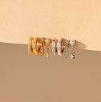 The Dainty Pave Earrings Set, featuring five pairs of gold and silver hoop earrings with gem and charm details, is displayed elegantly on a beige surface.