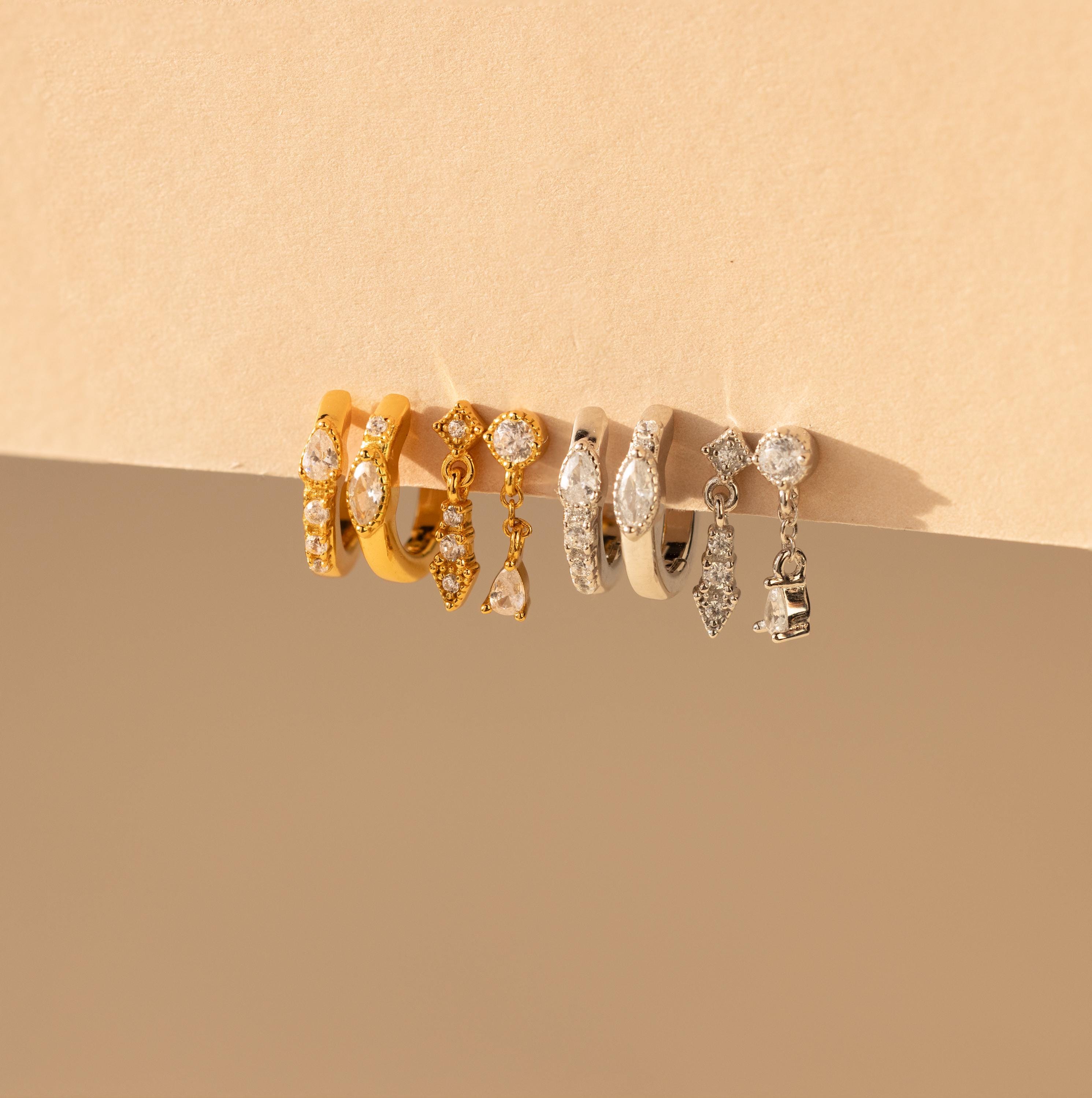 The Dainty Pave Earrings Set, featuring five pairs of gold and silver hoop earrings with gem and charm details, is displayed elegantly on a beige surface.