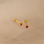 The Ruby Flatback Studs, featuring small red gemstones set in gold, rest on a light beige surface and add a subtle pop of color to your collection.