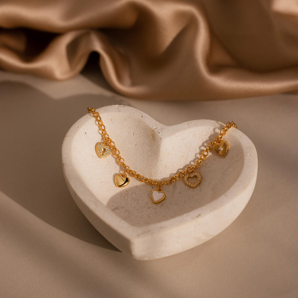 The Heart Charm Bracelet features delicate gold heart charms, beautifully displayed in a white heart-shaped dish on a soft beige fabric background—an elegant gift for your girlfriend.