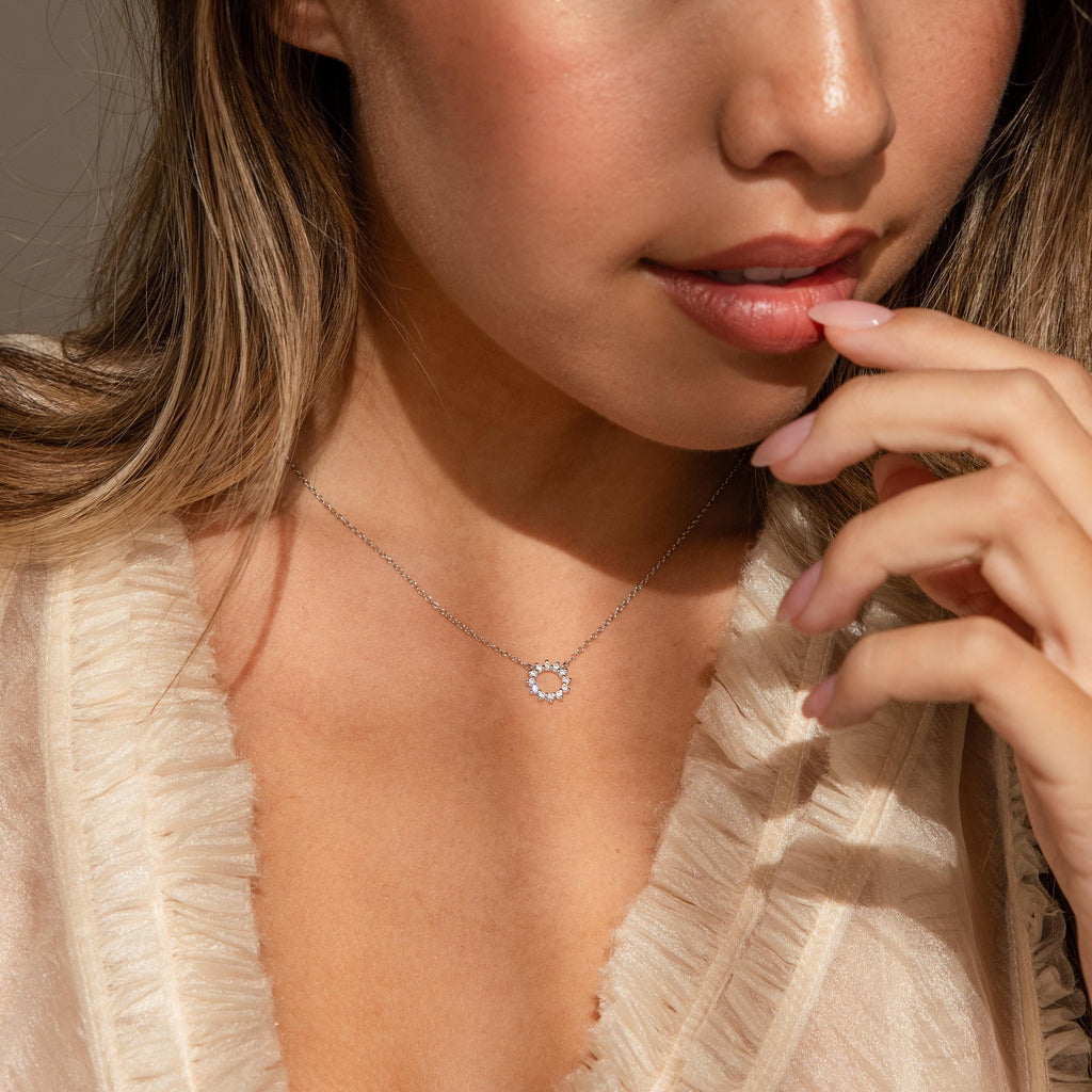 A woman touches her chin gently while wearing a ruffled cream blouse and the Diamond Sun Necklace, a delicate sunburst pendant.