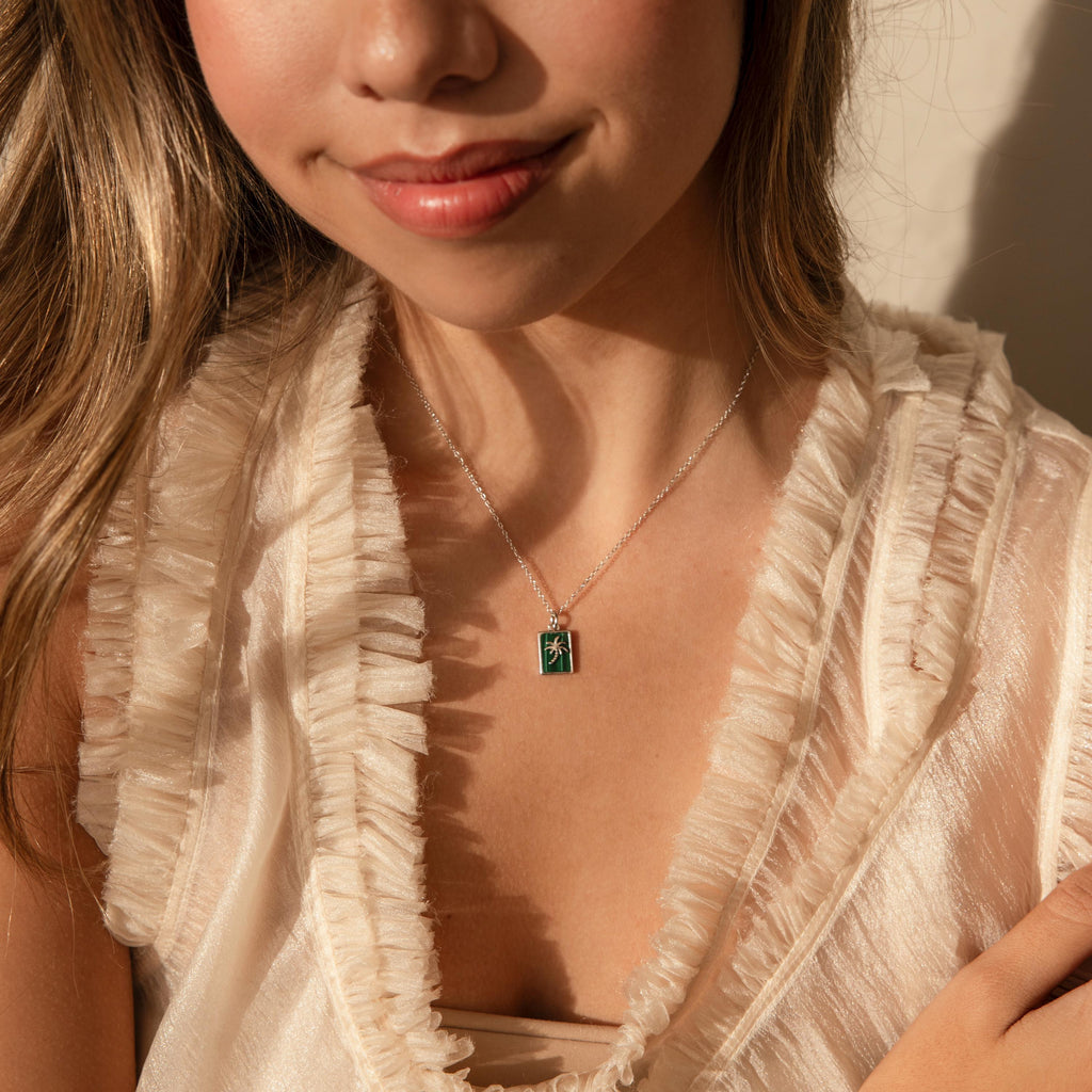 A woman wears a ruffled cream blouse and the Malachite Palm Tree Necklace, which features a green malachite gemstone pendant.