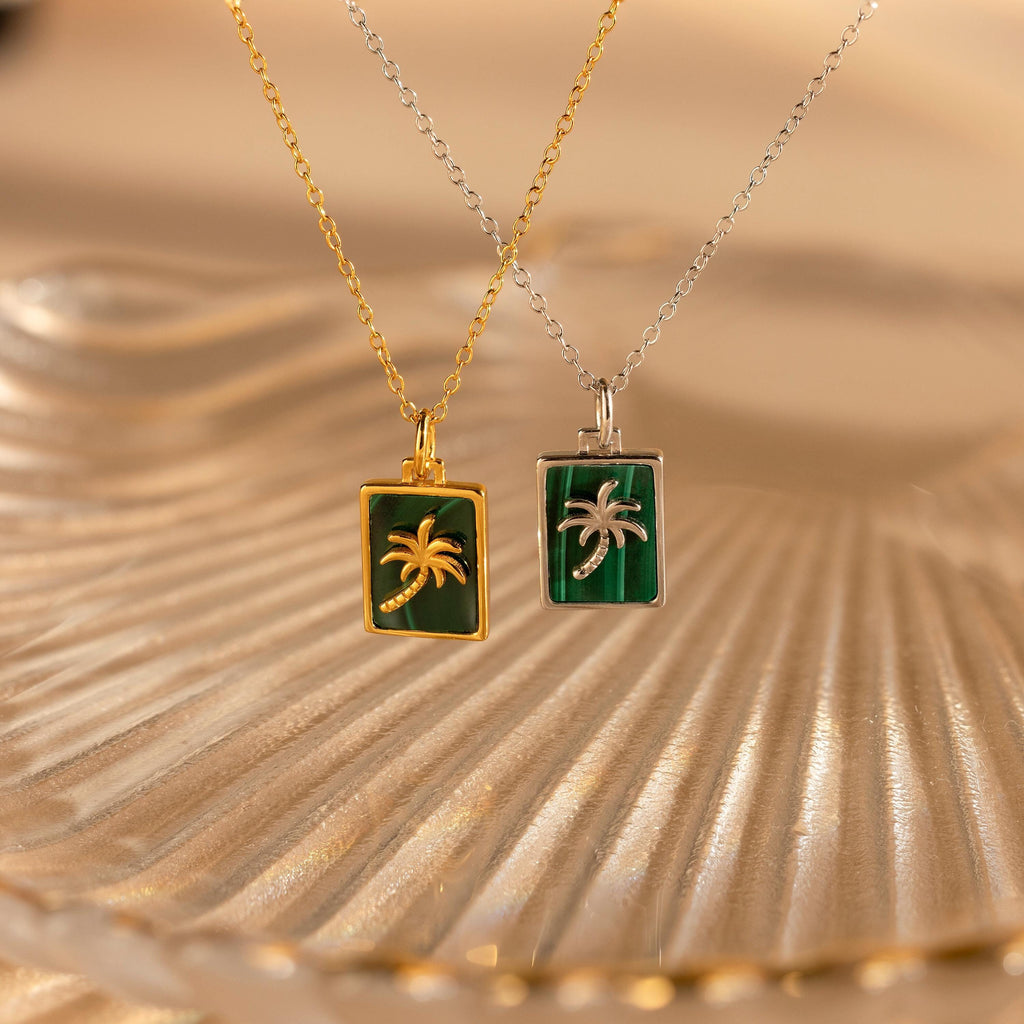 Two Malachite Palm Tree Necklaces—one gold and one silver— from our collection, displayed on textured glass.