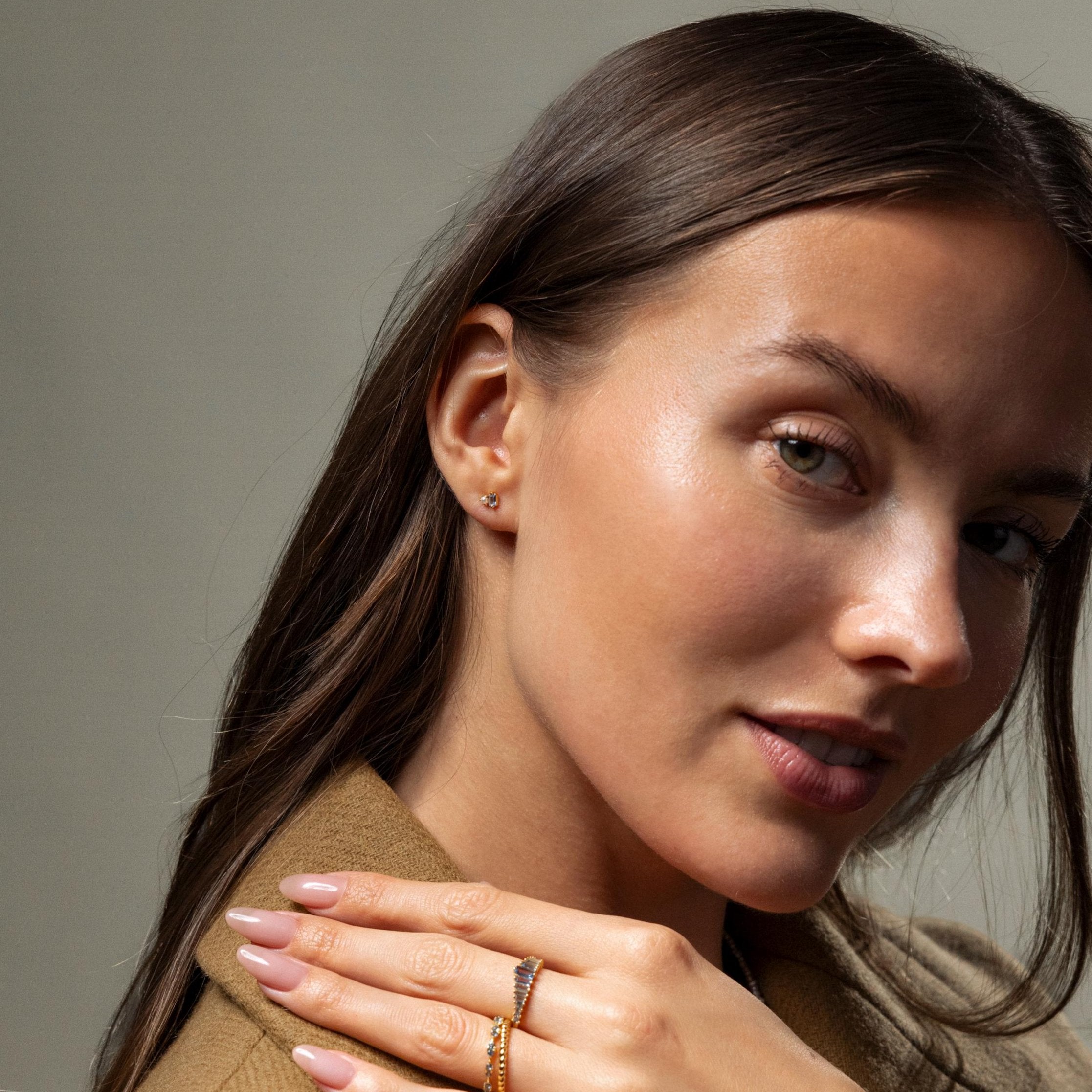 A woman with long brown hair in a tan coat and minimal jewelry poses with her hand near her face, highlighting the Pearl Aquamarine Studs that lend a subtle sparkle to her elegant style.