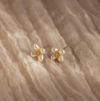 Pearl Flower Studs feature clusters of pearls and small gemstones in a floral design, beautifully displayed on textured beige fabric.