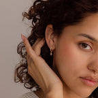 A person with curly hair touches their ear, highlighting the Pearl Ivy Flower Huggies—a small gold earring featuring a dangling charm.
