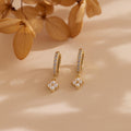 The Pearl Ivy Flower Huggies in 18K Gold feature pearl and crystal details, evoking a floral design. Displayed on a beige surface with dried flowers nearby.