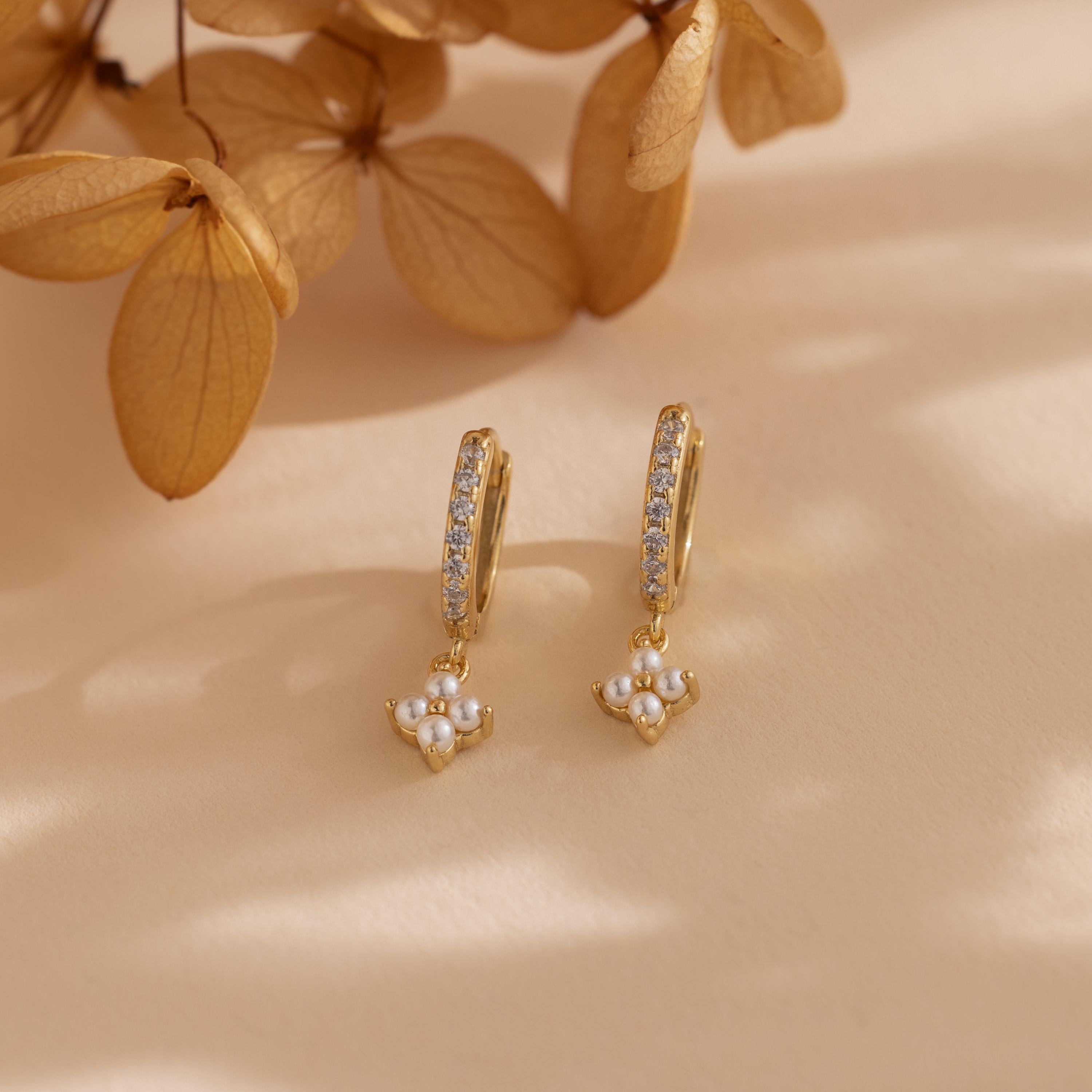 The Pearl Ivy Flower Huggies in 18K Gold feature pearl and crystal details, evoking a floral design. Displayed on a beige surface with dried flowers nearby.