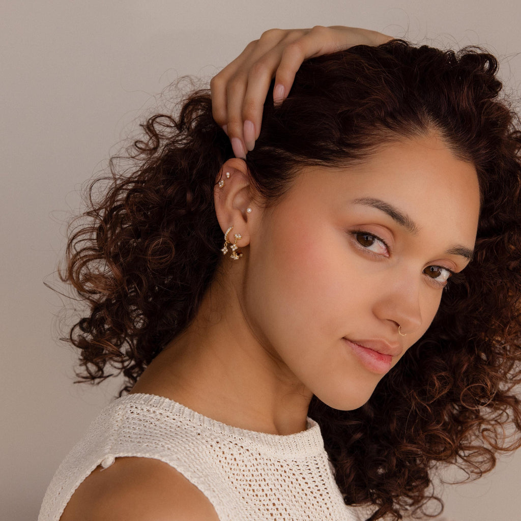 A woman with curly hair touches her ear, showcasing Pearl Ivy Flower Huggies, while wearing a cream knit top and looking at the camera.