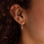 Close-up of an ear with Dangling Pearl Diamond Huggies in 18K Gold, featuring elegant hoops with two small dangling diamonds, against curly brown hair.