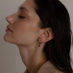 Woman in profile with closed eyes wears Black Diamond Drop Huggies—elegant hoop earrings featuring a small black bead—set against a neutral background.