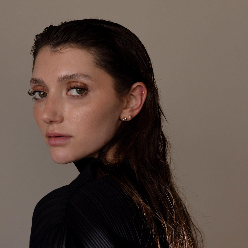 A woman with wet, dark hair in a black top looks over her shoulder against a plain background, showcasing Medium Diamond Bezel Flatback Studs that add a touch of elegance to her jewelry collection.