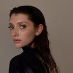 A woman with wet, dark hair in a black top looks over her shoulder against a plain background, showcasing Medium Diamond Bezel Flatback Studs that add a touch of elegance to her jewelry collection.