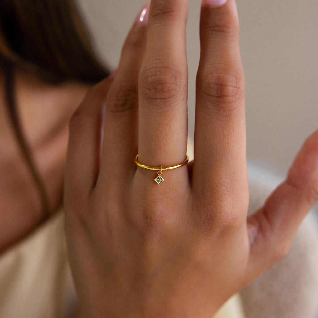 A hand wearing the Dainty Birthstone Drop Ring, featuring a small diamond-shaped charm on the ring finger—an ideal piece for personalized jewelry lovers.