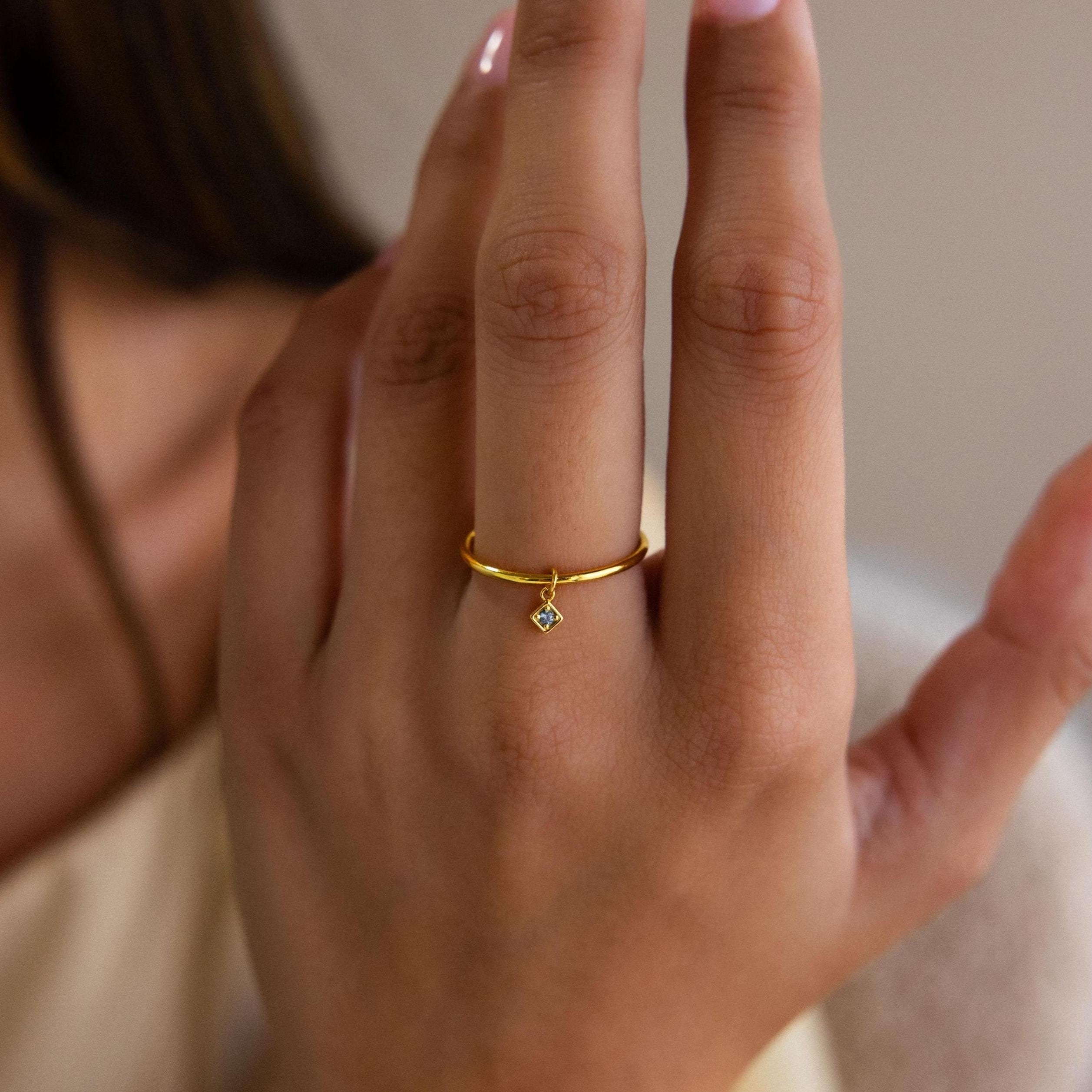 A hand wearing the Dainty Birthstone Drop Ring, featuring a small diamond-shaped charm on the ring finger—an ideal piece for personalized jewelry lovers.