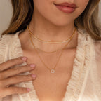 A woman wearing a cream ruffled blouse showcases the Diamond Sun Necklace, featuring a shimmering sunburst pendant, as she touches her collarbone with manicured fingers.