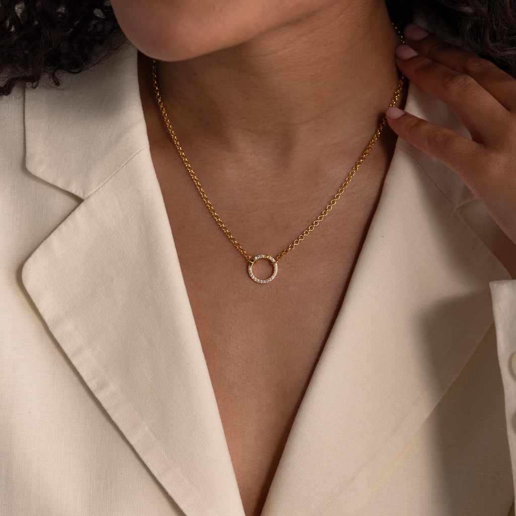 A woman wearing a cream blazer and a gold necklace featuring the Pave Diamond Charm Clip with a circular pendant and birthstone ring charm, hand touching her collar.