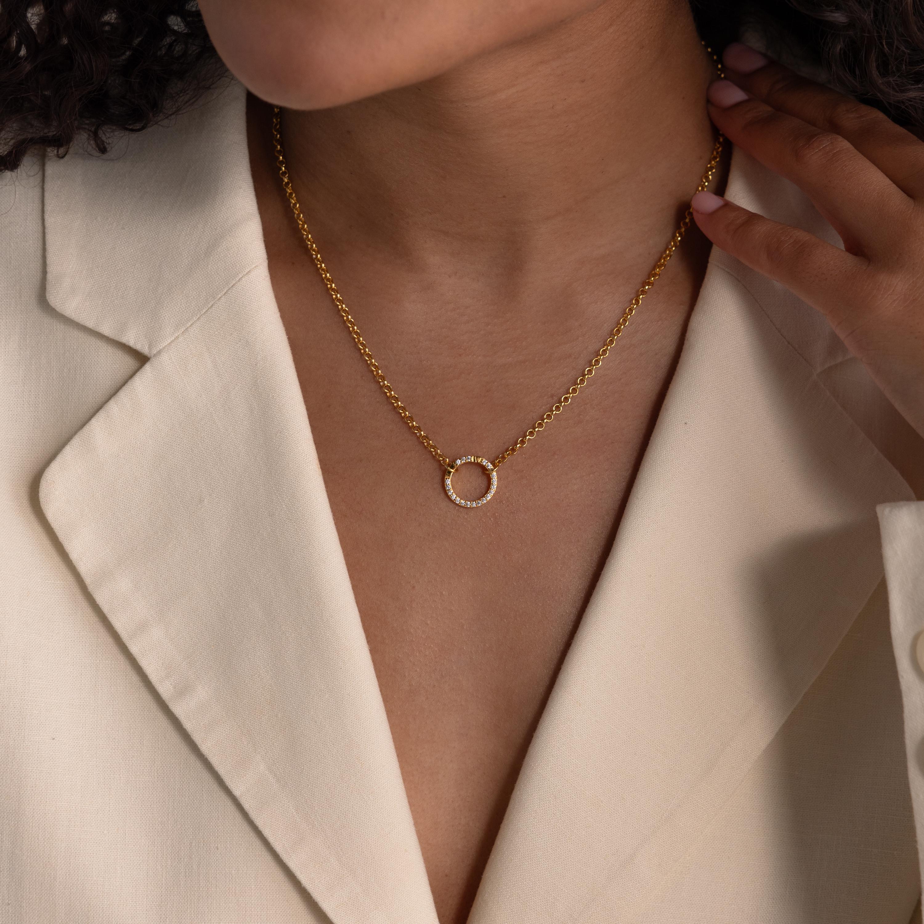 A woman wearing a cream blazer and a gold necklace featuring the Pave Diamond Charm Clip with a circular pendant and birthstone ring charm, hand touching her collar.